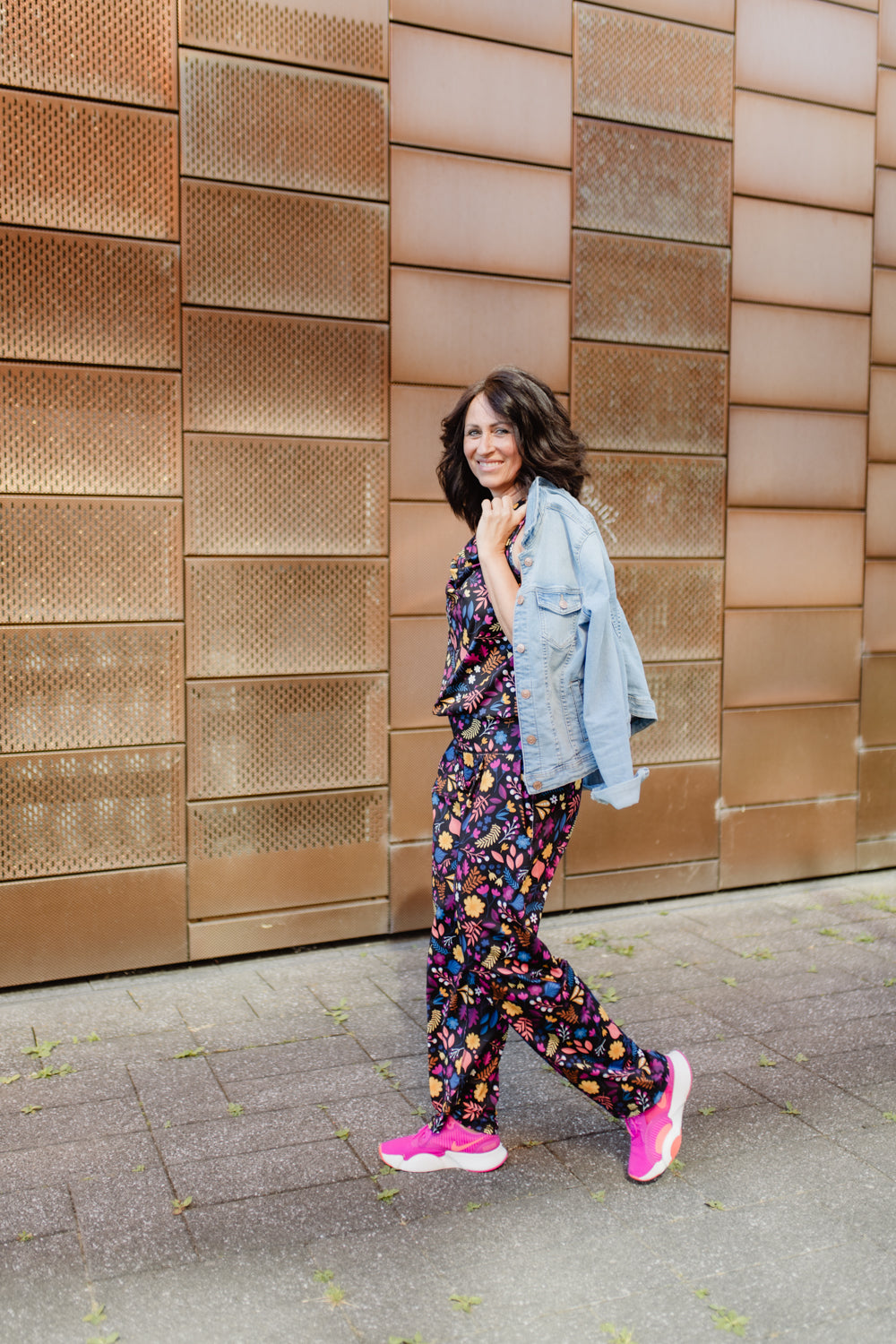Lächelnde brünette Frau mit Jumpsuit in Blumenmuster und rosa Turnschuhen, gehend durch die Stadt. Jeansjacke übergeworfen.