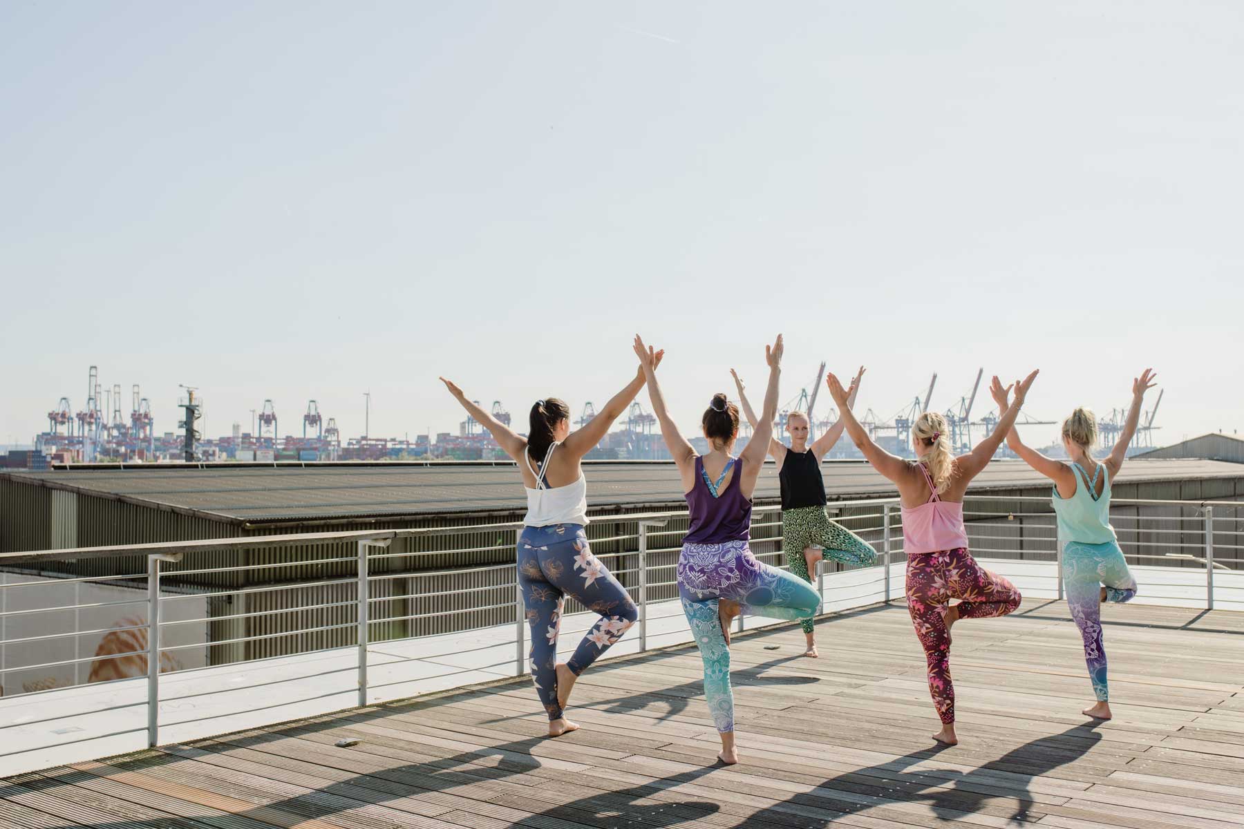 Fünf Frauen am Hafen in Yoga-Jumpsuits, welche Yoga-Übungen ausführen. Hände in der Luft, baruß. Einbeinige Yogaübung.