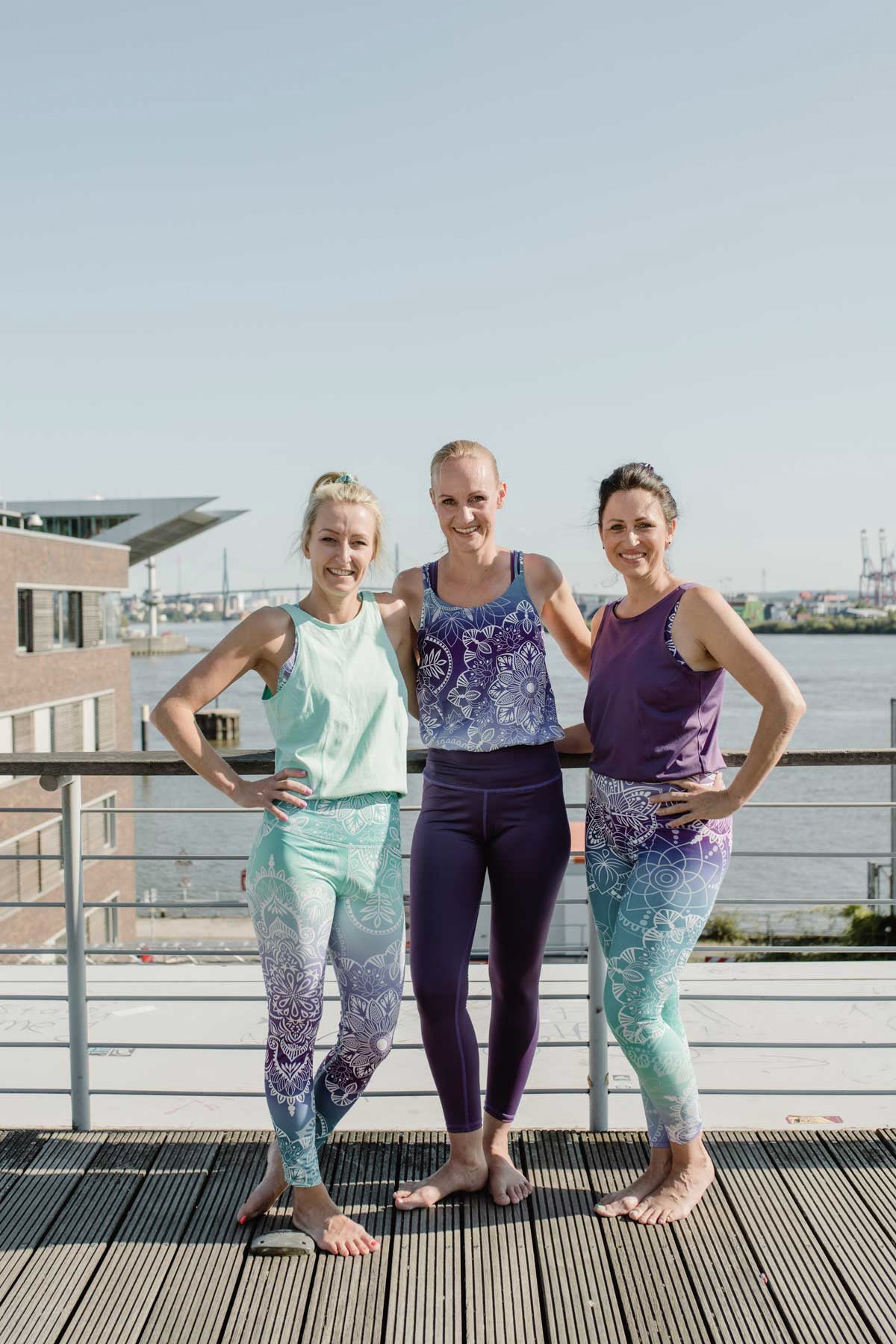 Drei lächelnde Frauen am Hafen mit lila und blauen Yoga Jumpsuits. Hände in die Seiten gestemmt, barfuß und lächelnd. Arm in Arm mit Freundinnen.