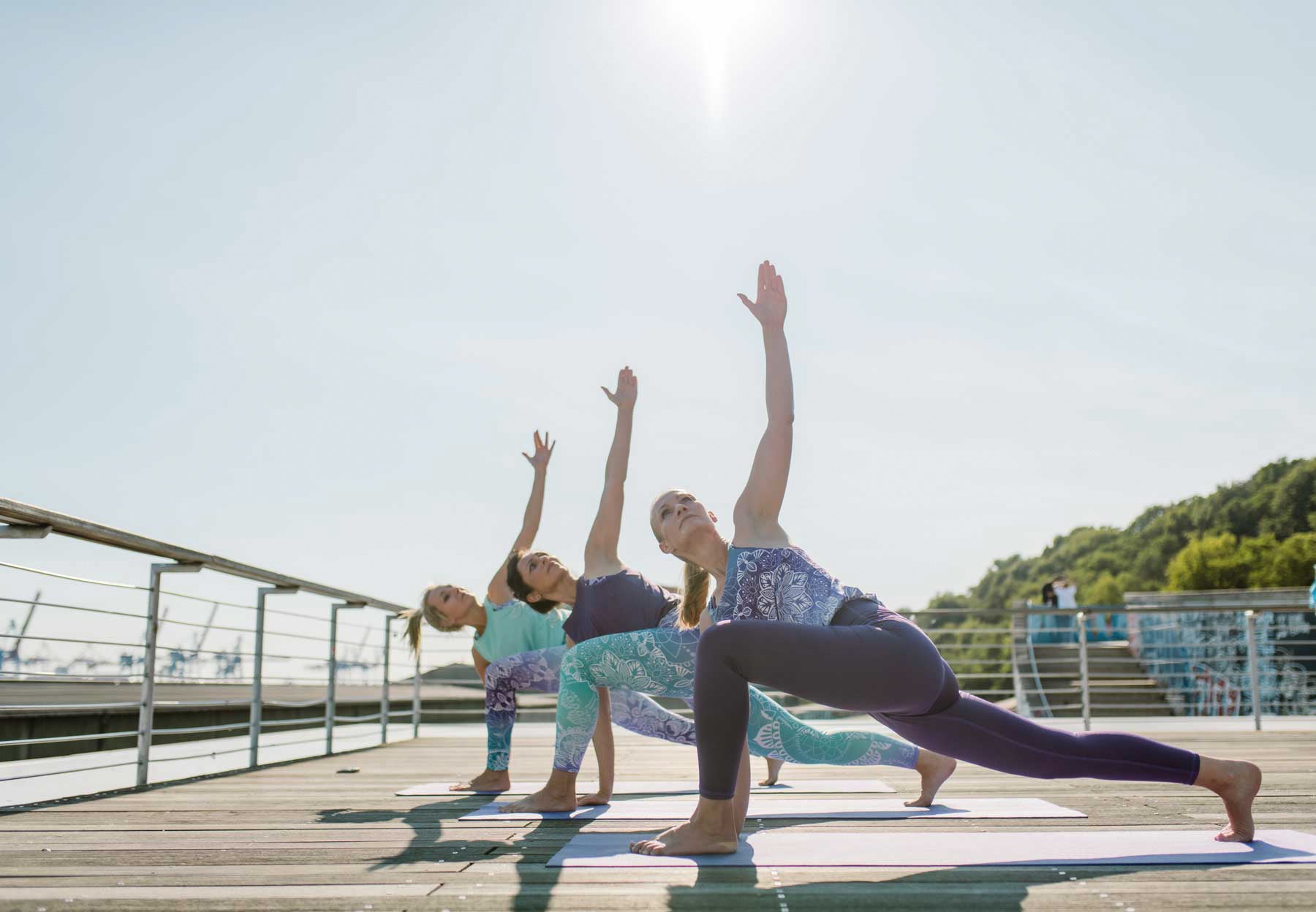 Drei Frauen in Yoga Jumpsuits auf Holzboden am Hafen in einer Yoga Pose. Barfuß, Arm in die Luft.