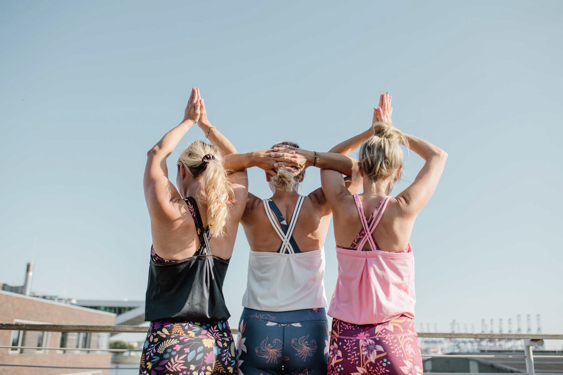 Drei Frauen in Yoga Jumpsuits von hinten betrachtet mit verschränkten Armen über dem Kopf. Unterschiedliche Farben der Jumpsuits.