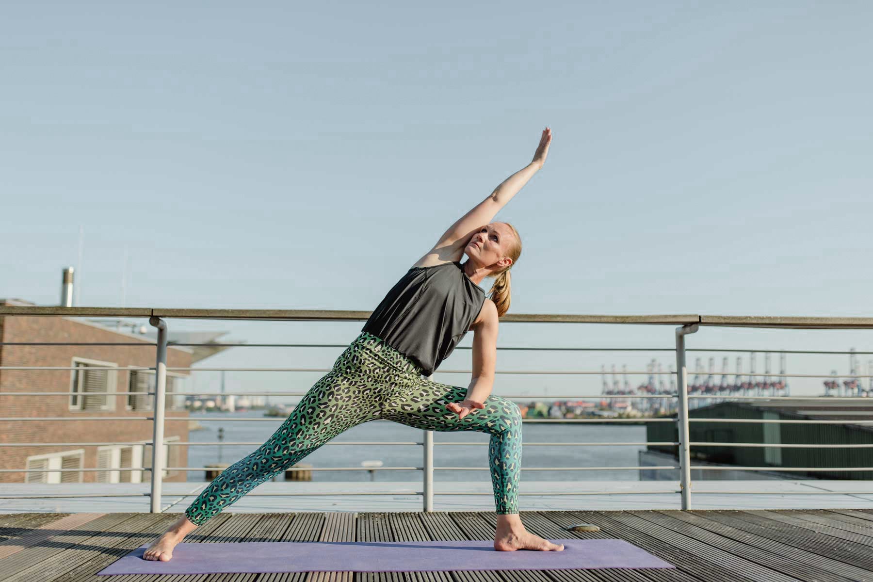 Jumpsuit in leo grünen Muster in Yoga Pose. Blondine in Yogahaltung barfuß auf Yogamatte am Hafen.