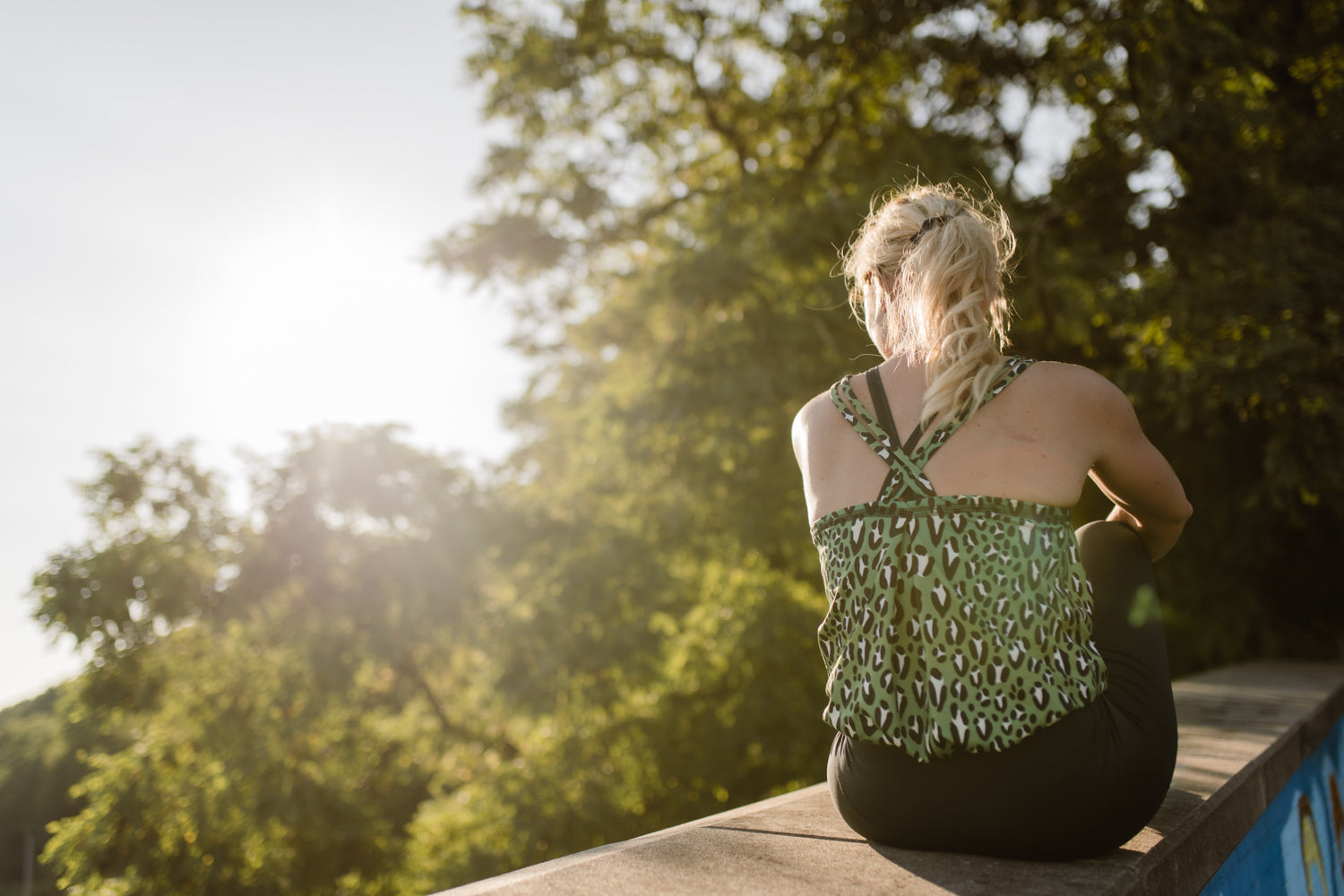 Blondine in grünem Yogajumpsuit sitzend auf einer Wand von hinten fotografiert mit Natur und Sonnenschein im Hintergrund