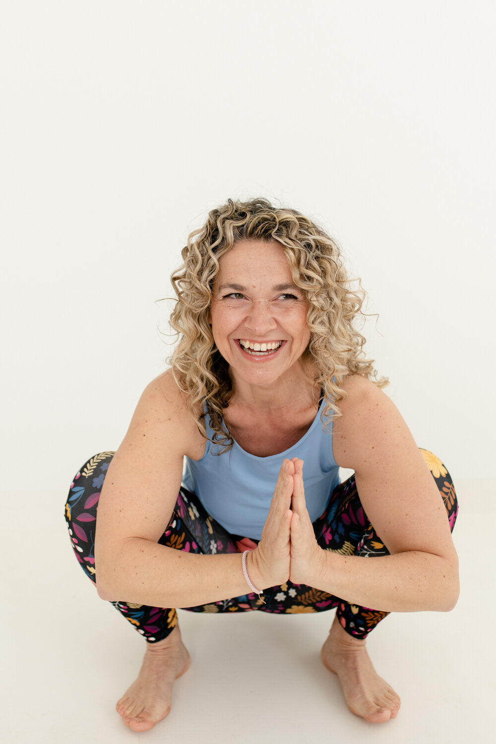 Lachende Frau mit blonden Locken barfuß in Froschpose mit hellblauem YogaJumpsuit Oberteil und Blumenmuster Unterteil