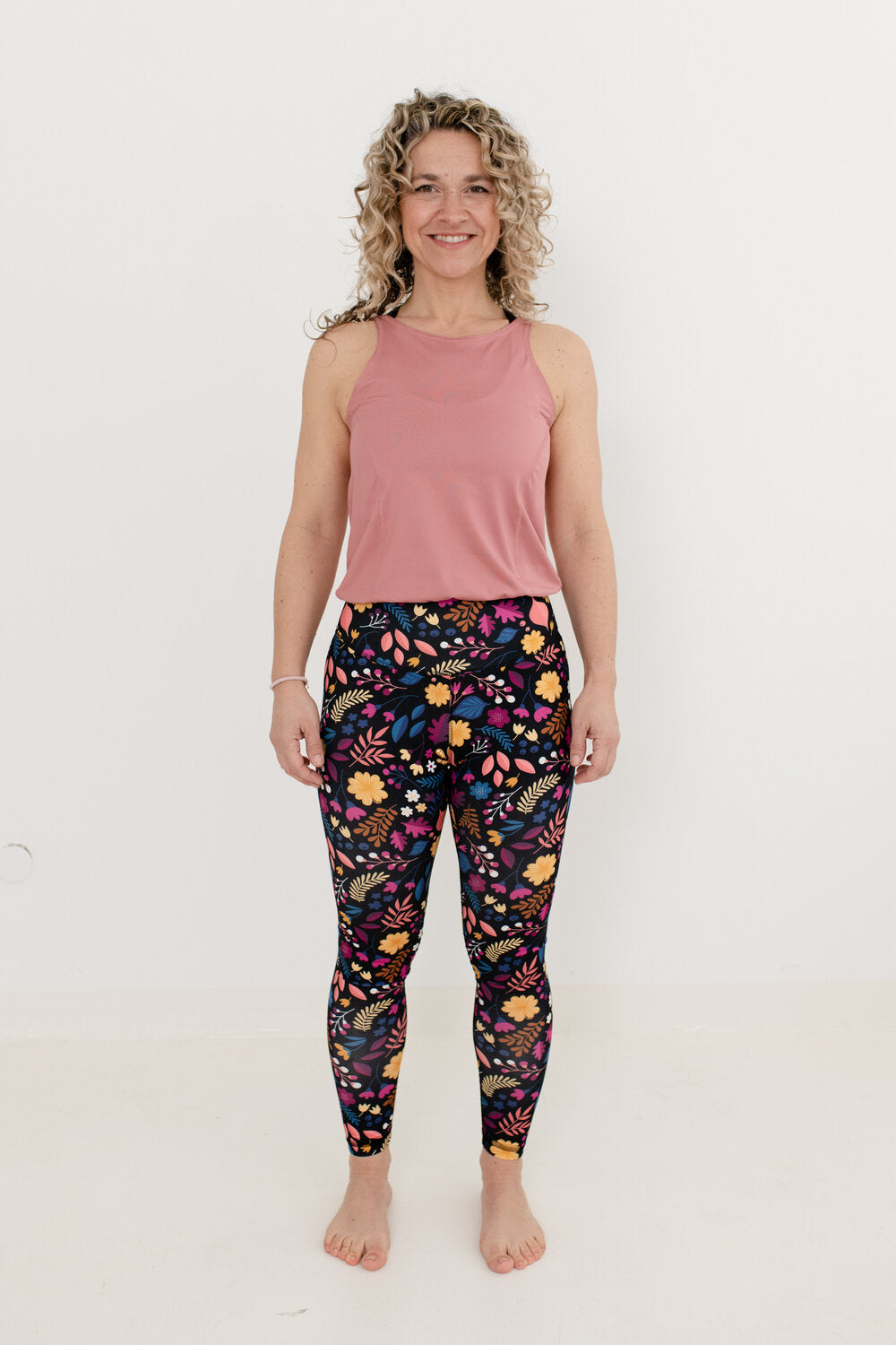 Lächelnde Blondine mit Locken trägt einen altrosa Yoga Jumpsuit mit Blumenmuster Unterteil