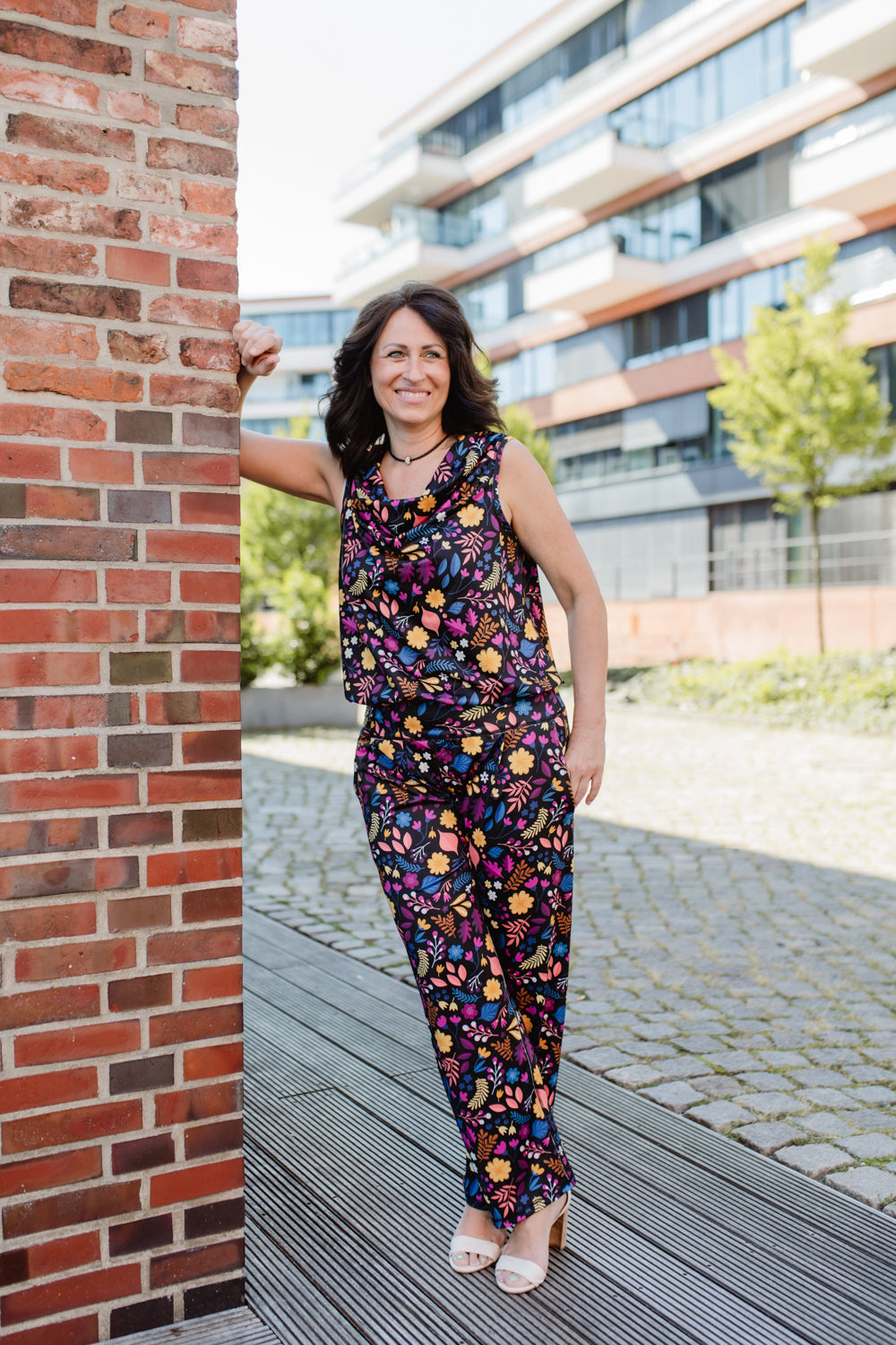 Lächelnde Brünette in Jumpsuit mit Blumenmuster, an eine Ziegelsteinwand gelehnt.